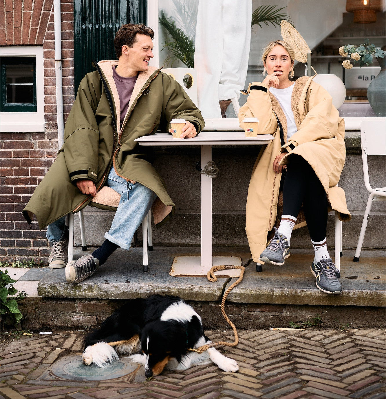 Two people sitting at a small outdoor cafe table in front of a shop window, each wearing a long oversized coat in olive green and beige, with a black, white, and brown dog lying on the brick pavement in the foreground.