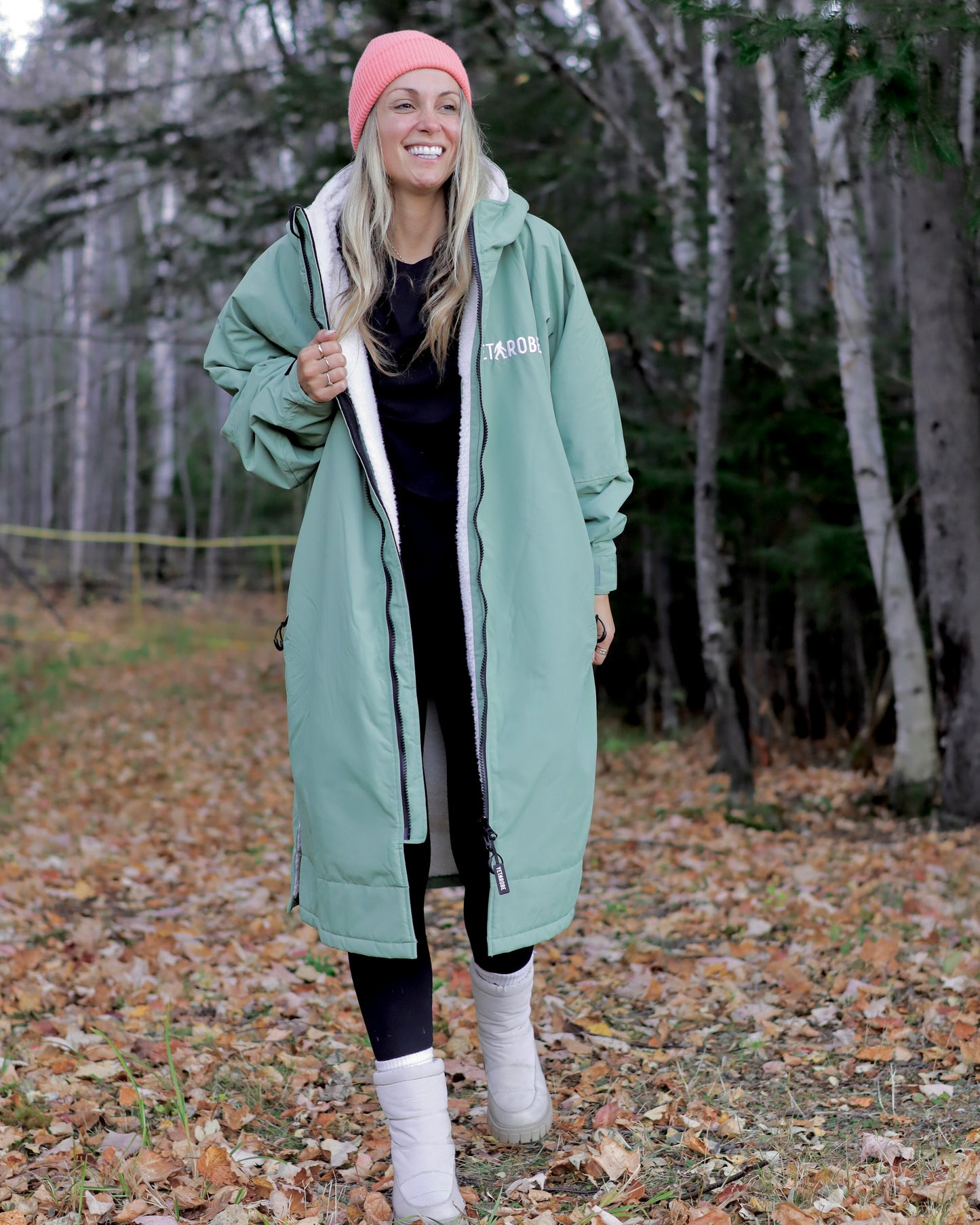 Woman wearing a long green coat with a brand logo, standing in a forest.